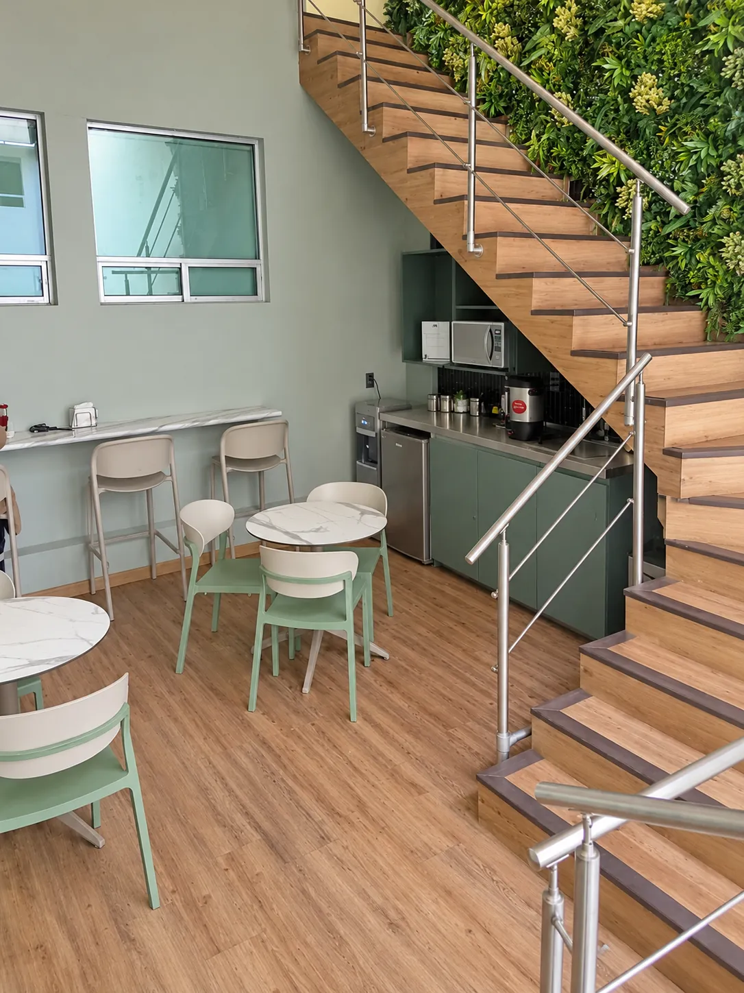 Cafetería bajo la escalera con muro de plantas, mesas y bancos, OfiRent Del Valle, Ciudad de México
